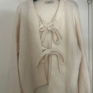 Krämvit kofta med knyt från Gina Tricot - Supermysig krämvit kofta från Gina Tricot med ribbstickat material och två stora knyt framtill. Koftan har lång ärm och en oversized känsla, perfekt att slänga på sig när det är kyligt. Enkel att styla till olika outfits.