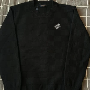 Stickad LV tröja - Svart stickad tröja märkt Louis Vuitton med  rutmönster i materialet och broderad vit text på bröstet.  Oanvänd och ny. Size M. Stor i storlek