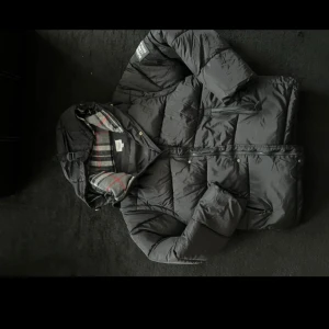 Svart pufferjacka Burberry M - Svart pufferjacka från Burberry London England i storlek M, med oversize passform. Jackan har huva med klassiskt rutig foder i grått, vitt och rött, samt dragkedja framtill och flera fickor. Perfekt för kalla dagar med sin quiltade design och snygga detaljer.