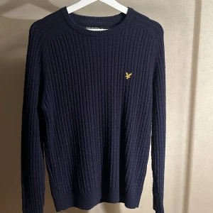 Mörkblå stickad tröja från Lyle & Scott - Säljer en mörkblå stickad tröja från Lyle & Scott med ribbat mönster och klassisk rund hals. Tröjan har lång ärm och den ikoniska gula loggan broderad på bröstet. Perfekt för kyliga dagar och enkel att matcha med jeans eller chinos.