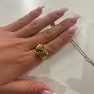 Guldfärgad ring med hjärtdesign - Cool guldfärgad ring med unik hjärtformad design och ett litet blixtformat snitt i mitten. Ringen har en bredare front och smalnar av baktill. Perfekt statement-piece för dig som gillar att sticka ut med accessoarer. Den är köpt för ungefär 300kr och har tydliga tecken på användning om man kollar nära. Det har lossnat färg på kanterna på ringen. Pris kan diskuteras❤️