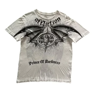 Rare Affliction Ozzy Osbourne t-shirt - Vit t-shirt från Affliction med stora svarta tryck fram och bak, inspirerad av Ozzy Osbourne. Framsidan har texten 'Prince Of Darkness' och en stor demon med vingar, baksidan har ett kors med vingar och Ozzy Osbourne-tryck. Klassisk passform och crewneck. Grym rockstil. RIP Ozzy Osbourne