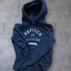 Mörkblå hoodie från Abercrombie & Fitch - Mysig mörkblå hoodie från Abercrombie & Fitch med tryck framtill. Klassisk huva med snörning och känguruficka. Perfekt för chill dagar och enkel att matcha med jeans eller joggers.