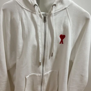 Vit hoodie med rött hjärta från Ami - Vit hoodie från Ami med dragkedja, huva med snörning och känguruficka. På bröstet finns ett rött hjärta med bokstaven A. Perfekt för en clean och stilren look. Passar L med