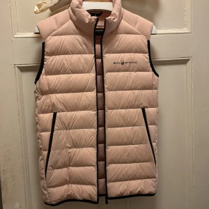 Ljusrosa väst från Sail Racing - Snygg ljusrosa väst från Sail Racing (helt oanvänd)  med svart dragkedja och svarta detaljer vid fickor och kanter. Västen har hög krage, två sidofickor med dragkedja och quiltad design. Perfekt för lager-på-lager och chill stil. Orginal priset var cirka 690 kr. Med övre inne ficka också.