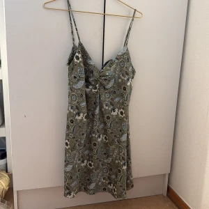 Grön blommig kortklänning med knyt - Snygg grön kortklänning med blommigt mönster och tunna axelband. Klänningen har en v-ringad front och ett knyt under bysten som ger en fin form. Perfekt för varma dagar och har en lätt och luftig känsla.