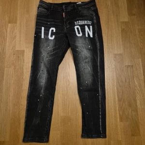 Svarta Dsquared2 ICON jeans med kedja - Snygga svarta jeans från Dsquared2 med vit 'ICON'-text framtill och coola slitningar samt färgstänk. Bakfickorna har en unik kedjedetalj och Dsquared2-logga i rött och vitt. Jeansen har en rak passform och är tillverkade i klassiskt denim-material.
