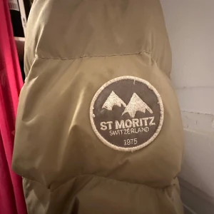 Beige pufferjacka St Moritz - Säljer en beige pufferjacka från St Moritz med broderad patch på ärmen. Jackan har hög krage, snörning i nederkant och praktiska fickor. Materialet är syntet och jackan är quiltad för extra värme. Perfekt för kalla dagar.