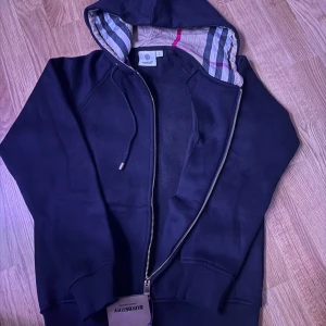 Svart hoodie från Burberry med dragkedja - Svart hoodie från Burberry med klassiskt rutigt foder i huvan och snygga metalldetaljer på snörena. Tröjan har hel dragkedja framtill och ribbade muddar. Perfekt för dig som vill ha en stilren och lyxig look med ikoniska Burberry-detaljer.