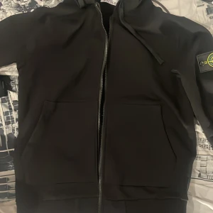 Svart hoodie från Stone Island - Svart hoodie från Stone Island med klassisk logga på ärmen. Tröjan har dragkedja framtill, två stora fickor och huva med snörning. Tillverkad i mjuk bomull, perfekt för en clean och stilren look.