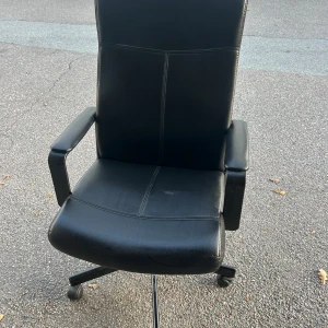 Kontorsstol - Detta är en svart kontorsstol med hög rygg, armstöd och hjul för enkel förflyttning. Stolen har viss ytligt slitage på sitsen men är i överlag gott skick. Perfekt för hemmakontoret eller arbetsplatsen. 