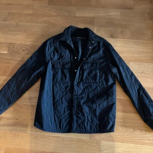 Svart jacka  J.Lindeberg - Säljer en svart quiltad overshirt från J.Lindeberg. Jackan har två bröstfickor med lock, knappar framtill och lång ärm. Perfekt för lager-på-lager och har en clean, stilren look som funkar till det mesta.