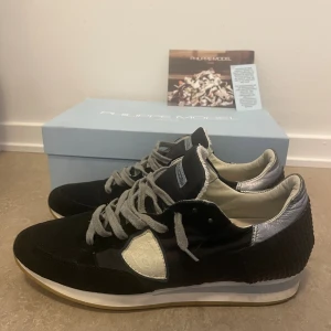 Philippe Model marinblå sneakers - Blå sneakers från Philippe Model med grå snörning, silverdetaljer vid hälen och vit logotyp på sidan. Ovandelen är i en mix av mocka och syntet, med vit och beige sula. Snygg och sportig design som passar till många outfits. Endast skorna medföljer