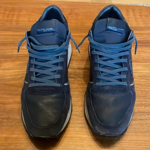 Mörkblå sneakers från Philippe Model - Snygga mörkblå sneakers från Philippe Model med ovandel i skinn och textil. Skorna har rund tå, platt sula och klassisk snörning. Snygga detaljer i olika blå nyanser. Perfekta för dig som gillar stilrena och bekväma sneakers.