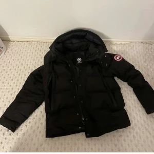 Svart dunjacka från Canada Goose - Säljer en svart dunjacka från Canada Goose med huva och klassisk logga på ärmen. Jackan har dragkedja och knappar framtill, samt två stora fickor. Det finns också både päls och box.