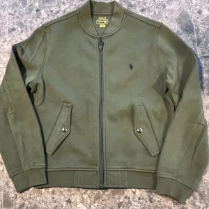 Polo Ralph Lauren jacka+byxa - Snygg olivgrön jacka och byxa från Polo Ralph Lauren. Jackan har dragkedja framtill, två fickor med lock och knapp samt klassisk logga broderad på bröstet. Storlek M (10-12 år).