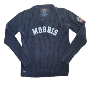 Mörkblå ribbad tröja Morris M - Snygg mörkblå ribbstickad tröja från Morris med vit broderad logga på bröstet och patch på ärmen. Tröjan har rund hals och långa ärmar, perfekt för dig som gillar klassisk stil med en sportig touch.