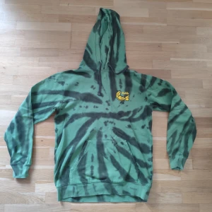 Grön tie-dye hoodie från Vans - Säljer en grön och svart tie-dye hoodie från Vans med stor gul peace-logga och texten 'Vans Off The Wall The Peaceful Outdoors' på ryggen. Framtill finns en mindre logga och klassisk känguruficka. Perfekt för dig som gillar streetwear och coola mönster.