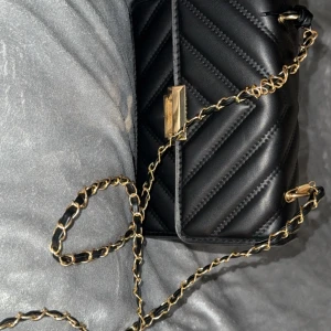 Svart quiltad axelväska med guldkedja - Snygg svart axelväska i quiltad design med guldfärgad kedja och låsdetalj. Väskan har ett stilrent mönster i diagonala ränder och är tillverkad i skinnimitation. Perfekt storlek för det viktigaste och ger en lyxig känsla till din outfit.