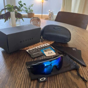 Oakley sportglasögon blå lins - Säljer ett par Oakley sportglasögon med cool blå spegellins och svart båge. Modellen har en omslutande form som sitter grymt bra vid träning och utomhusaktiviteter. Kommer med originalfodral, putsduk och box. Perfekt för dig som vill ha stil och funktion.