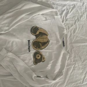 Vit Palm Angels sweatshirt med björn - Vit sweatshirt från Palm Angels med ikoniskt björnmotiv i beige och brunt på bröstet. Tröjan har rund halsringning, långa ärmar och broderad Palm Angels-logga under motivet. Perfekt statement piece för streetwear-stilen.
