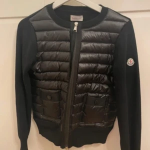 Svart pufferjacka från Moncler - Svart pufferjacka från Moncler med quiltad framsida och två fickor med tryckknappar. Jackan har rund halsringning, långärmade stickade ärmar och dragkedja framtill. Moncler-logga på ärmen och diskreta detaljer som ger en clean look. 