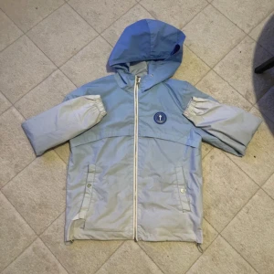 Trapstar blå vindjacka - Säljer en blå och ljusblå vindjacka från Trapstar med huva och dragkedja. Jackan har stort Trapstar-tryck på ryggen och rund blå patch med T-logga på bröstet. Två fickor framtill med knappstängning och elastiska muddar vid ärmslut. Perfekt för dig som gillar streetwear.