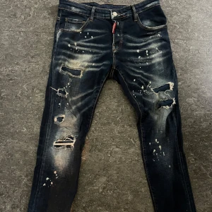 Dsquared2 mörkblå slitna jeans - Riktigt snygga mörkblå jeans från Dsquared2 med coola slitningar, hål och färgstänk. Slim fit-modell med klassisk femficksdesign och tydliga detaljer. Jeansen är tillverkade i Italien och har en modern streetkänsla. 