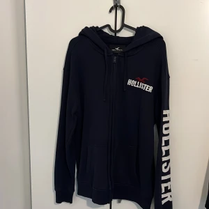 Mörkblå hoodie från Hollister M - Mörkblå hoodie från Hollister med dragkedja, broderad logga på bröstet och stort tryck på ärmen. Tröjan har huva med snörning och två fickor framtill. Insidan är mjuk och fleecefodrad för extra komfort.