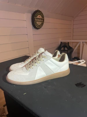 Maison Margiela -  Sneakers  - Maison Margiela sneakers i vitt med beige detaljer och gummisula. Klassisk låg modell med rund tå, snörning och paneler i mocka och skinn. Tungan har den ikoniska Margiela-etiketten med siffror. Perfekt för dig som gillar minimalistisk och clean stil.