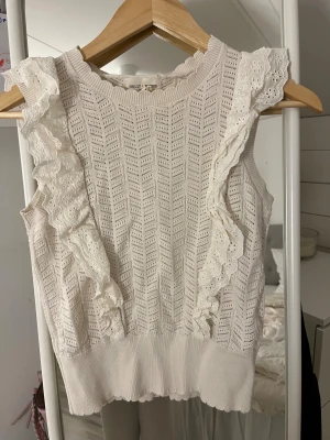 Vit ärmlös topp med volang från Lindex  - Supersöt vit topp från Lindex med broderade volanger längs axlarna. Stickad struktur med hålmönster och ribbad kant nedtill. Perfekt för dig som gillar romantiska detaljer och ljusa färger. Rund halsringning och ärmlös design.