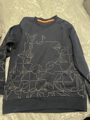  Louis Vuitton sweatshirt med broderi - Svart sweatshirt från Louis Vuitton med unikt broderat mönster i beige tråd på framsidan och ärmarna. Mönstret består av LV-logga, noter och blommor. Rund hals och långärmad modell, med orange etikett i nacken. Perfekt för dig som gillar exklusiva streetwear-vibes.