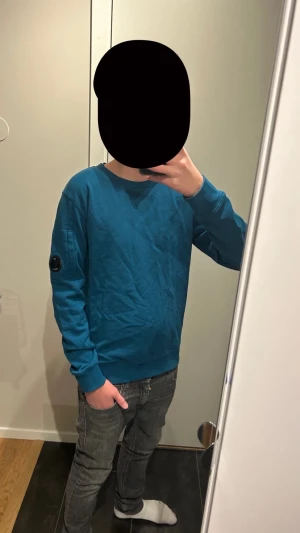 Blå crewneck från C.P. Company - Säljer en blå crewneck från C.P. Company med deras ikoniska lins på vänster ärm. Tröjan har rund hals, ribbade muddar och är i mjuk bomull. Perfekt för dig som gillar stilrena och snygga plagg med en twist den är 14 år men sitter som Xs köpt på Nk.  