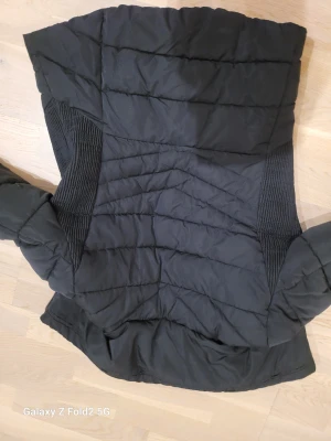 Svart pufferjacka från Zara - Snygg svart pufferjacka från Zara med mjuk fuskpäls i kragen och guldfärgade dragkedjor. Jackan har quiltad design, ribbade paneler på sidorna och två fickor med dragkedja. Perfekt för kalla dagar och har en modern, stilren look.