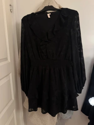 Svart kortklänning med volanger - Supersnygg svart kortklänning från Nelly med volanger och v-ringning. Klänningen har långärmade transparenta ärmar och ett subtilt blommigt mönster i tyget. Smockad midja ger en skön passform och extra volym i kjolen.