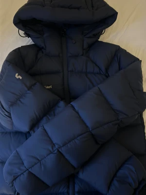 Mörkblå pufferjacka Peak Performance XS - Säljer en mörkblå pufferjacka från Peak Performance i storlek XS. Jackan har huva, dragkedja framtill och är vadderad för extra värme. Perfekt för kalla dagar och har en clean, sportig look. Materialet är syntet och dunblandning.