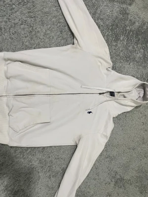 Vit hoodie från Polo Ralph Lauren - Vit hoodie från Polo Ralph Lauren i custom fit med dragkedja framtill och två fickor. Klassisk broderad logga i marinblått på bröstet och snörning i huvan. Mjuk bomullskvalitet och ribbade muddar vid ärmslut och nederkant. Perfekt för chill dagar.