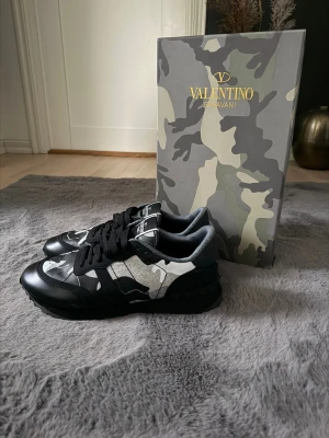 Valentino Garavani Rockrunner sneakers - Snygga Valentino Garavani Rockrunner sneakers i svart, grått och vitt med coolt camouflage-mönster. Skorna har snörning, platt sula med grovt mönster och rund tå. Materialet är en mix av skinn och textil. Kommer med originalkartong, dustbag och extra skosnören.