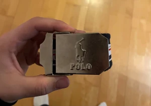 Mörkblått Polo bälte med metallspänne - Snyggt mörkblått bälte från Polo med ett robust metallspänne där Polo-loggan är ingraverad. Bältet har ett randigt band i mörkblått, vitt och rött med Polo-tryck och logga längs hela längden. Bältet är 120cm långt. Aldrig använt. Perfekt accessoar för att lyfta din outfit.