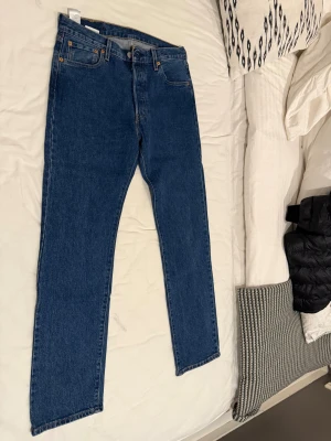 Levi's 501 blå jeans straight fit - Klassiska Levi's 501 jeans i blå denim med raka ben och normal passform. Byxorna har fem fickor, knappgylf och den ikoniska läderpatchen bak i midjan. Perfekta för dig som gillar tidlös stil och vill ha ett par jeans som funkar till allt.använd 1 gång 