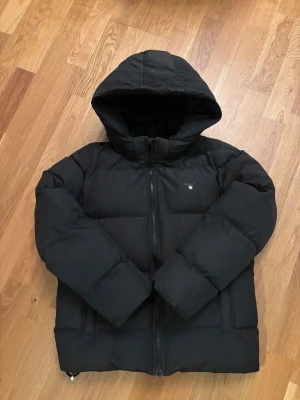 Svart pufferjacka från GANT med huva - Svart pufferjacka från GANT med huva och dragkedja framtill. Jackan har ett stilrent GANT-logo på bröstet och är vadderad för extra värme. Passar dig som gillar en enkel och clean look under kalla dagar.