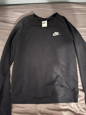 Svart Nike sweatshirt med logga - Svart sweatshirt från Nike med klassisk vit broderad logga på bröstet. Tröjan har rund hals, långärmade ärmar och är gjord i mjuk bomullsmix. Perfekt för en chill och sportig stil. Passar dig som gillar enkel och clean design.