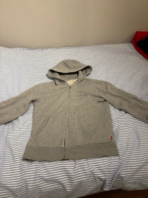 Grå hoodie från Levi's med dragkedja - Grå hoodie från Levi's i storlek S med dragkedja framtill och två fickor. Klassisk design med broderad Levi's-logga på bröstet och mjukt fleecefoder på insidan. Perfekt för kyliga dagar och enkel att matcha med jeans eller joggers.