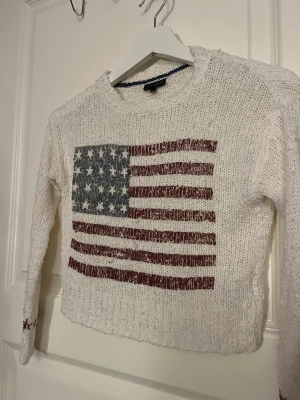Vit stickad tröja med USA-flagga - Vit stickad tröja från Bondelid med stort tryck av amerikanska flaggan på bröstet och röda stjärnor på ärmsluten. Tröjan har grovstickad struktur, rund hals och långa ärmar. Perfekt för dig som gillar statement-plagg och vill sticka ut.