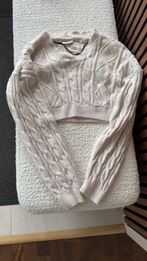 Beige croppad stickad tröja Noisy May - Croppad stickad tröja från Noisy May i beige med flätstickat mönster och v-ringning. Tröjan har långa ärmar och ribbade muddar, perfekt för lager-på-lager. Snygg och trendig modell som passar till höst och vinter.