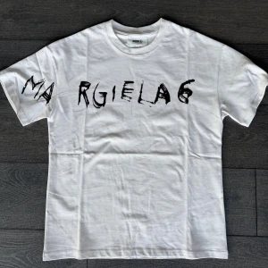 Maison Margiela t shirt  - Snygg vit t-shirt från Maison Margiela MM6 med svart, handskriven logotyp över bröstet och ärmen. Klassisk rund hals och korta ärmar. Tillverkad i mjuk bomull för skön känsla och stilren look.