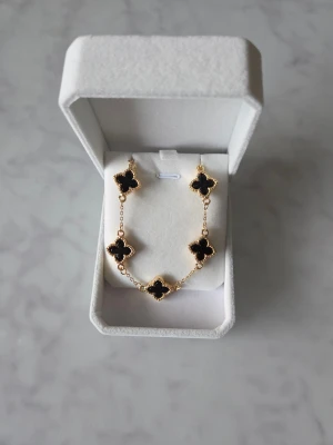 Guldarmband med svarta fyrklöver - Snyggt armband i guldton med fem svarta fyrklöverformade hängen. Kedjan är tunn och har justerbar längd med små ringar och en diskret platta vid låset. Perfekt för dig som gillar stilrena detaljer med en twist.