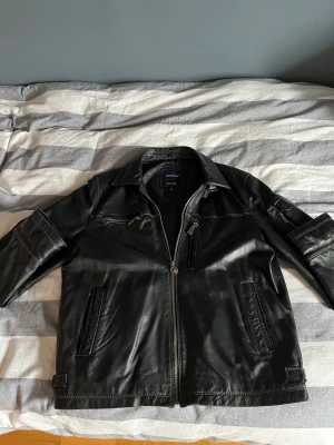 Svart skinnjacka Petroff Silver XL - Svart skinnjacka från Petroff Silver i storlek XL. Jackan har flera dragkedjor, fickor och snygga detaljer med kontrastsömmar. Klassisk krage och coola spännen vid ärmslutet ger en edgy vibe. Perfekt för dig som gillar stilren och tuff look.