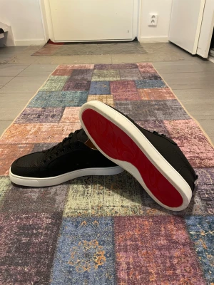 Christian Louboutin - Svarta Sneakers - Svarta sneakers från Christian Louboutin i exklusivt skinn med klassisk röd sula. Skorna har rund tå, svart snörning och vit platt sula. Insidan är beige och materialet är strukturerat för en lyxig känsla. Perfekta för dig som vill sticka ut med en ikonisk detalj. Skorna är i mycket bra skick och har endast en liten repa på sidan som syns på sista bilden. Dom här skorna är även svåra att hitta 🐦‍🔥❤️