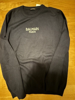 Mörkblå stickad tröja Balmain Paris - Snygg mörkblå stickad tröja från Balmain Paris med vit broderad logga på bröstet. Rund halsringning och långa ärmar med ribbade muddar. Perfekt för dig som gillar stilrena designer och exklusiva märken.